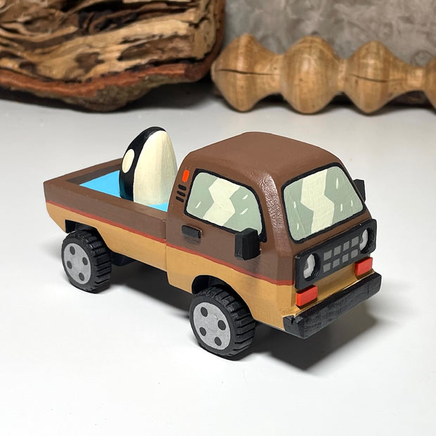 Painted small whittled wooden sculpture of a brown striped pick up truck, with the pick up filled with water. The head of a killer whale peeks out from the water.