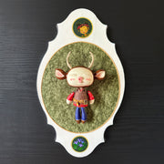 Ceramic doll mounted to a wooden plaque with painted elements. Doll has a oval shaped, closed eye face with kissy lips. It has antlers, deer ears and is dressed like an outdoorsman, with a puffy brown utility vest and jeans.