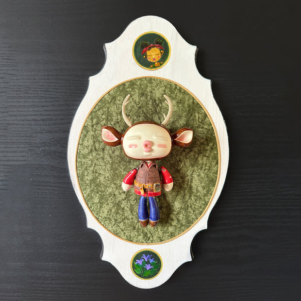 Ceramic doll mounted to a wooden plaque with painted elements. Doll has a oval shaped, closed eye face with kissy lips. It has antlers, deer ears and is dressed like an outdoorsman, with a puffy brown utility vest and jeans.