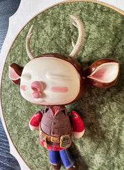 Ceramic doll mounted to a wooden plaque with painted elements. Doll has a oval shaped, closed eye face with kissy lips. It has antlers, deer ears and is dressed like an outdoorsman, with a puffy brown utility vest and jeans.