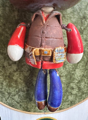 Ceramic doll mounted to a wooden plaque with painted elements. Doll has a oval shaped, closed eye face with kissy lips. It has antlers, deer ears and is dressed like an outdoorsman, with a puffy brown utility vest and jeans.