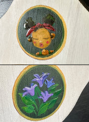 Close up of painted elements on the plaque: a girl with closed eyes and her lips pursed, and a set of purple flowers.