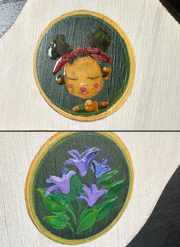 Close up of painted elements on the plaque: a girl with closed eyes and her lips pursed, and a set of purple flowers.