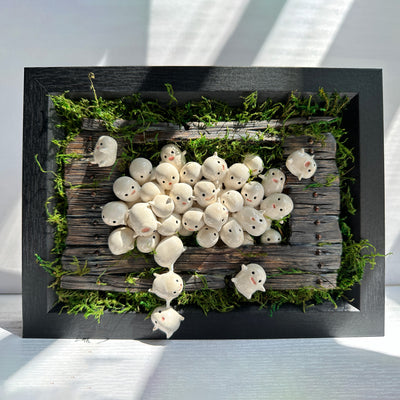 Mixed media sculpture of many small cute white blob creatures emerging from wooden floor boards with a large hole in the middle and greenery all around. Piece is within a black shadowbox frame without glass.