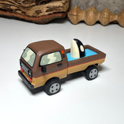 Painted small whittled wooden sculpture of a brown striped pick up truck, with the pick up filled with water. The head of a killer whale peeks out from the water.