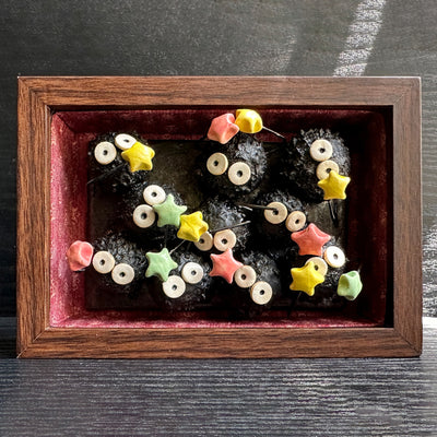 Diorama style sculptural work of many black soot sprites holding colorful stars. They're within a wooden box with red fabric lining it.