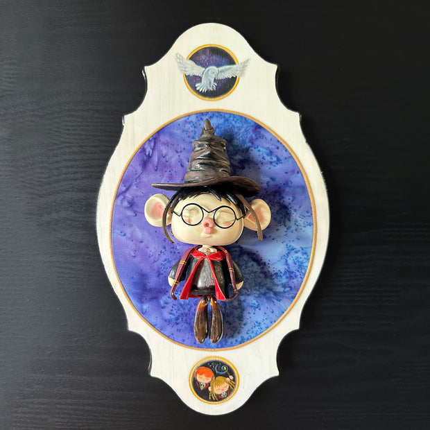 Ceramic doll mounted to a wooden plaque with painted elements. Doll is designed like Harry Potter with an oval shaped head, closed eye face with kissy lips. He wears a brown wizard hat and black and red cloak.