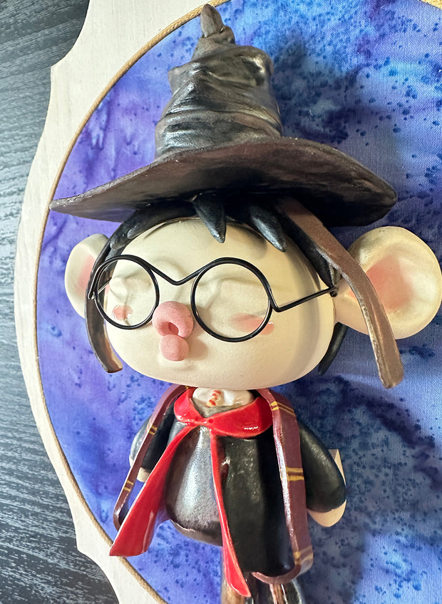 Ceramic doll mounted to a wooden plaque with painted elements. Doll is designed like Harry Potter with an oval shaped head, closed eye face with kissy lips. He wears a brown wizard hat and black and red cloak.