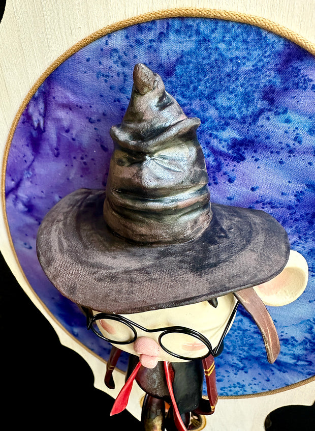 Ceramic doll mounted to a wooden plaque with painted elements. Doll is designed like Harry Potter with an oval shaped head, closed eye face with kissy lips. He wears a brown wizard hat and black and red cloak.