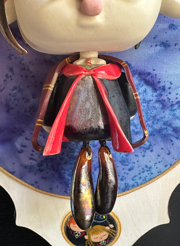 Ceramic doll mounted to a wooden plaque with painted elements. Doll is designed like Harry Potter with an oval shaped head, closed eye face with kissy lips. He wears a brown wizard hat and black and red cloak.