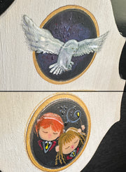 Close up of 2 painted details on the plaque: a white snowy owl mid flight and Ron Weasly and Hermoine wearing their school uniforms, positioned closely together holding a wand.