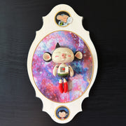 Ceramic doll mounted to a wooden plaque with painted elements. Doll is designed like Astroboy with an oval shaped head, closed eye face with kissy lips. His chest is open, revealing a circuit board and a heart.