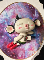 Ceramic doll mounted to a wooden plaque with painted elements. Doll is designed like Astroboy with an oval shaped head, closed eye face with kissy lips. His chest is open, revealing a circuit board and a heart.