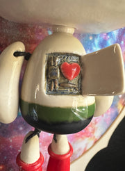 Ceramic doll mounted to a wooden plaque with painted elements. Doll is designed like Astroboy with an oval shaped head, closed eye face with kissy lips. His chest is open, revealing a circuit board and a heart.