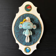 Ceramic doll mounted to a wooden plaque with painted elements. Doll is designed like a smurf with an oval shaped head, closed eye face with kissy lips. Atop its floppy white hat is a yellow flower.