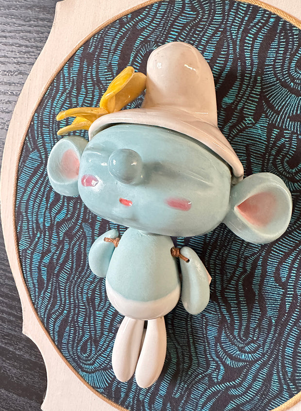 Ceramic doll mounted to a wooden plaque with painted elements. Doll is designed like a smurf with an oval shaped head, closed eye face with kissy lips. Atop its floppy white hat is a yellow flower.