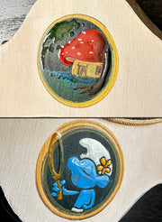 Close up of 2 painted details: one is a mushroom house in the woods and the other is of a smurf with a large magnifying glass or bubble blower.