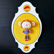 Ceramic doll mounted to a wooden plaque with painted elements. Doll is designed like Chucky from Child's Play with an oval shaped head, closed eye face with kissy lips. It wears a striped shirt, jean overalls and has red mid length hair.