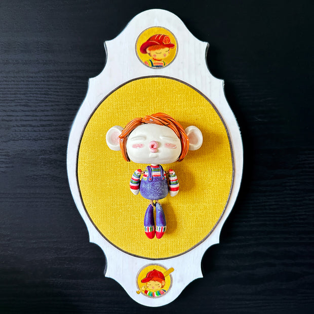 Ceramic doll mounted to a wooden plaque with painted elements. Doll is designed like Chucky from Child's Play with an oval shaped head, closed eye face with kissy lips. It wears a striped shirt, jean overalls and has red mid length hair.