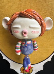 Ceramic doll mounted to a wooden plaque with painted elements. Doll is designed like Chucky from Child's Play with an oval shaped head, closed eye face with kissy lips. It wears a striped shirt, jean overalls and has red mid length hair.