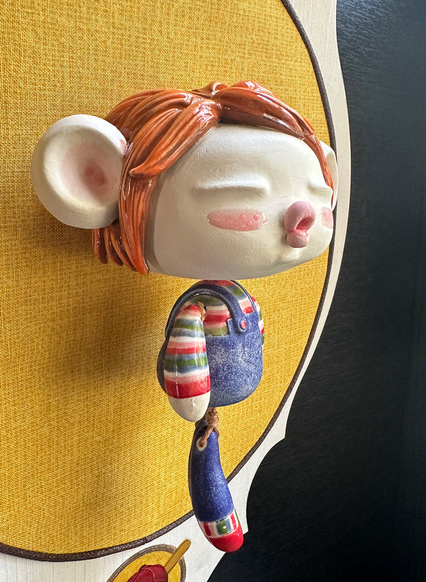 Ceramic doll mounted to a wooden plaque with painted elements. Doll is designed like Chucky from Child's Play with an oval shaped head, closed eye face with kissy lips. It wears a striped shirt, jean overalls and has red mid length hair.