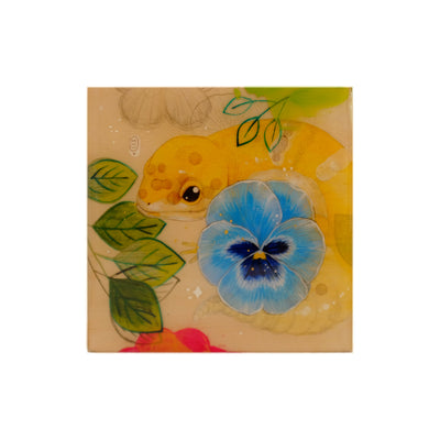 Painting on exposed wooden panel of a small yellow gecko, wrapped around itself and behind a blue pansy flower. Leaves of varying transparencies fill the piece.