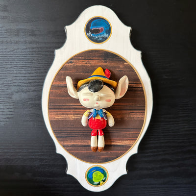 Ceramic doll mounted to a wooden plaque with painted elements. Doll is designed like Pinnochio with an oval shaped head, closed eye face with kissy lips.