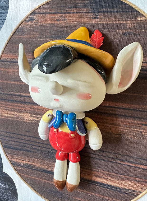 Ceramic doll mounted to a wooden plaque with painted elements. Doll is designed like Pinnochio with an oval shaped head, closed eye face with kissy lips.