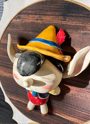 Ceramic doll mounted to a wooden plaque with painted elements. Doll is designed like Pinnochio with an oval shaped head, closed eye face with kissy lips.