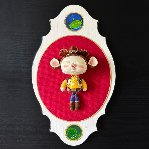 Ceramic doll mounted to a wooden plaque with painted elements. Doll is designed like Woody from Toy Story with an oval shaped head, closed eye face with kissy lips. He wears his standard outfit: yellow shirt with cow patterned vest, and a sheriff's hat. 