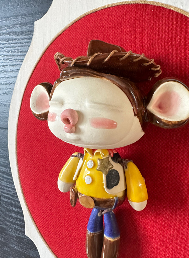 Ceramic doll mounted to a wooden plaque with painted elements. Doll is designed like Woody from Toy Story with an oval shaped head, closed eye face with kissy lips. He wears his standard outfit: yellow shirt with cow patterned vest, and a sheriff's hat. 