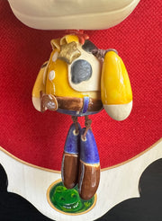 Ceramic doll mounted to a wooden plaque with painted elements. Doll is designed like Woody from Toy Story with an oval shaped head, closed eye face with kissy lips. He wears his standard outfit: yellow shirt with cow patterned vest, and a sheriff's hat. 