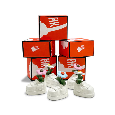 5 sculpture of white shoes, with single flowers growing out of each of them. Behind them are a stack of orange and white boxes, like small shoe boxes.