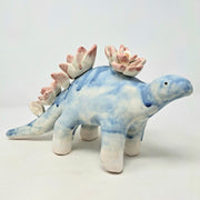 Ceramic sculpture of a blue dinosaur, likely a stegosaurus, with blue drip pattern body and pink drip pattern lotuses on its back in place of spikes.