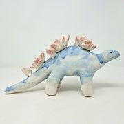 Ceramic sculpture of a blue dinosaur, likely a stegosaurus, with blue drip pattern body and pink drip pattern lotuses on its back in place of spikes.