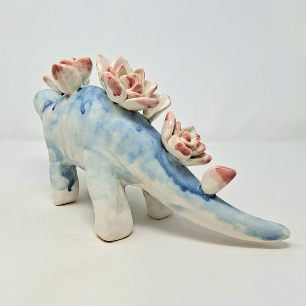 Ceramic sculpture of a blue dinosaur, likely a stegosaurus, with blue drip pattern body and pink drip pattern lotuses on its back in place of spikes.