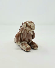 Ceramic sculpture of a tan and darker brown kaiju creature, sitting on the ground with its legs out in front of it. It has rounded spikes along its back.