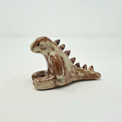 Ceramic sculpture of a tan and darker brown kaiju creature, sitting on the ground with its legs out in front of it. It has rounded spikes along its back.