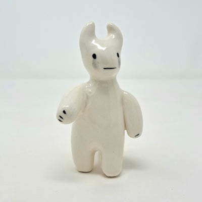 White glazed ceramic sculpture of a simplistic character standing on 2 legs, with its arms out at its side and 2 horns atop its head. It has a simple face, 2 dots for eyes and a line smile with tears below its eyes.