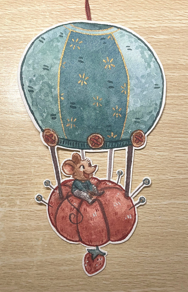 Die cut illustration on paper of a hot air balloon, with the basket portion a tomato shaped pin cushion. Riding atop it is a small mouse, smiling.