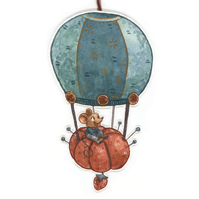 Die cut illustration on paper of a hot air balloon, with the basket portion a tomato shaped pin cushion. Riding atop it is a small mouse, smiling.