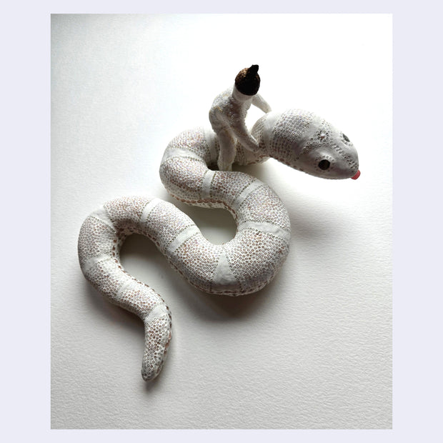 Sculpture of a white snake, seen from overhead. It has delicate patterning on its body making up its scales with thin white stripes. Riding on its neck is a woman with her arms loosely wrapped around it.