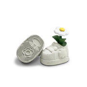 Sculpture of a small chunky white shoe with a single white flower growing from a green stem within it. The underside of the shoe is also show, with a circular flower design.