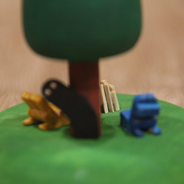 Sculptural piece of a round tree on a mound of grass. In front are 2 wooden chairs and an unlit bonfire. Behind the tree is a shadowy figure, peeking out.