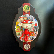 Sculpture of a doll with a wide head, closed eyes and its lips pursed in a kiss. It has large ears and wears a red robot helmet and has a red robot body with the limbs dangling. Its attached to a plaque with a collage of Giant Robot magazine covers.