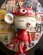 Sculpture of a doll with a wide head, closed eyes and its lips pursed in a kiss. It has large ears and wears a red robot helmet and has a red robot body with the limbs dangling. Its attached to a plaque with a collage of Giant Robot magazine covers.