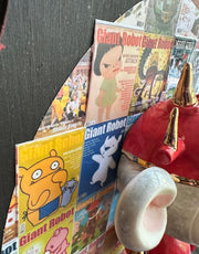 Sculpture of a doll with a wide head, closed eyes and its lips pursed in a kiss. It has large ears and wears a red robot helmet and has a red robot body with the limbs dangling. Its attached to a plaque with a collage of Giant Robot magazine covers.