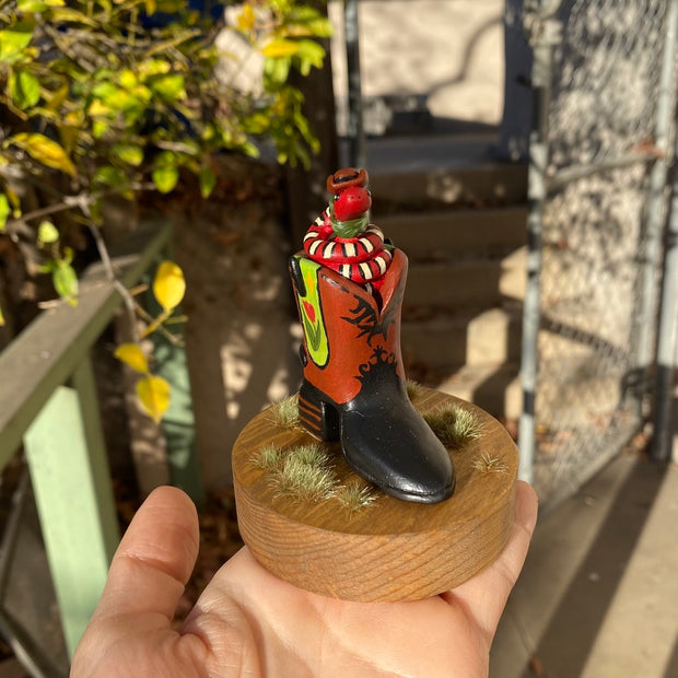 Sculpture of a red snake with black and white stripes, coiled up inside of a brown and black cowboy boot with a spur. Cowboy boot has a design element of a pink tulip with yellow stars on green background on each side of the boot. Snake wears a small cowboy hat and bandana. 