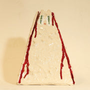 Offwhite ceramic sculpture of a pyramid shaped vase, with a wide base and a slightly narrow top. The vase is bumpy in texture and has a subtle cat face on the top. It has streaks of red running down the side of it.