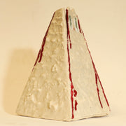 Offwhite ceramic sculpture of a pyramid shaped vase, with a wide base and a slightly narrow top. The vase is bumpy in texture and has a subtle cat face on the top. It has streaks of red running down the side of it.